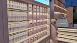 A well-organized display at a store, featuring shelves stocked with canned goods and neatly arranged boxes, primarily in soft pink and white colors. There are also a counter with a coffee machine and a digital screen displaying an image of a drink.