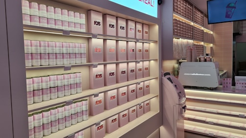 A well-organized display at a store, featuring shelves stocked with canned goods and neatly arranged boxes, primarily in soft pink and white colors. There are also a counter with a coffee machine and a digital screen displaying an image of a drink.