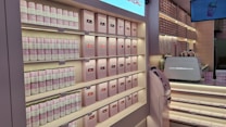 A well-organized display at a store, featuring shelves stocked with canned goods and neatly arranged boxes, primarily in soft pink and white colors. There are also a counter with a coffee machine and a digital screen displaying an image of a drink.