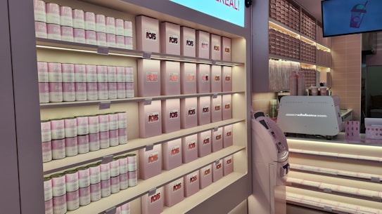 A well-organized display at a store, featuring shelves stocked with canned goods and neatly arranged boxes, primarily in soft pink and white colors. There are also a counter with a coffee machine and a digital screen displaying an image of a drink.