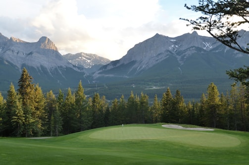 A lush, green golf course with a smooth putting green in the foreground, surrounded by tall evergreen trees. Majestic, rugged mountains loom in the background under a partly cloudy sky.