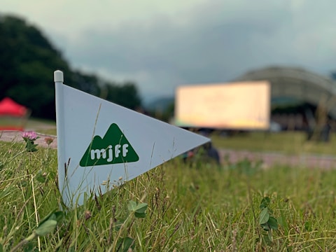 A triangular flag with a green mountain logo and the letters 'mjff' is positioned among grass, with a blurred background that includes a tent-like structure and a large screen. The scene suggests an outdoor event or festival.