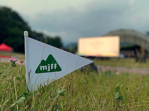 A triangular flag with a green mountain logo and the letters 'mjff' is positioned among grass, with a blurred background that includes a tent-like structure and a large screen. The scene suggests an outdoor event or festival.