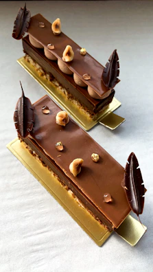 Two chocolate desserts with layered textures are artfully arranged on a rectangular golden plate. The top surface is glossy with chocolate decorations and garnished with hazelnuts and small, shiny edible decorative elements. The desserts are elongated and appear delicate, with chocolate leaves on each end.