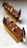 Two chocolate desserts with layered textures are artfully arranged on a rectangular golden plate. The top surface is glossy with chocolate decorations and garnished with hazelnuts and small, shiny edible decorative elements. The desserts are elongated and appear delicate, with chocolate leaves on each end.