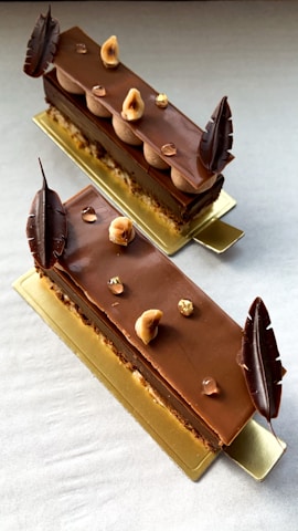 Two chocolate desserts with layered textures are artfully arranged on a rectangular golden plate. The top surface is glossy with chocolate decorations and garnished with hazelnuts and small, shiny edible decorative elements. The desserts are elongated and appear delicate, with chocolate leaves on each end.