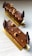 Two chocolate desserts with layered textures are artfully arranged on a rectangular golden plate. The top surface is glossy with chocolate decorations and garnished with hazelnuts and small, shiny edible decorative elements. The desserts are elongated and appear delicate, with chocolate leaves on each end.