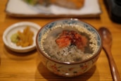 A bowl of rice soup with roasted salmon, garnished with seaweed and crunchy pearls. The bowl is ornately decorated and placed on a wooden surface. Nearby, there is a small plate with pickles and wasabi, and a wooden spoon lies beside the bowl.