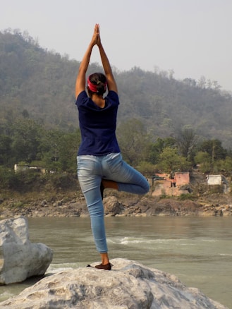A person is practicing yoga in a tree pose on a rock near a river, surrounded by forested hills in the background. The sky is overcast, creating a serene and peaceful setting.