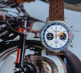 A wristwatch with a leather strap is placed on a magazine page that features an image of a motorcycle. The watch face is white with orange and blue hands and numerals, and has two small dials indicating seconds and minutes. The scene suggests a blend of adventure and style.