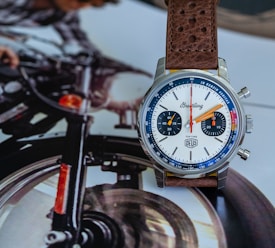 A wristwatch with a leather strap is placed on a magazine page that features an image of a motorcycle. The watch face is white with orange and blue hands and numerals, and has two small dials indicating seconds and minutes. The scene suggests a blend of adventure and style.