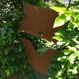 A rusted metal sculpture with abstract shapes is partially obscured by lush green foliage. The sculpture consists of two primary pieces, one with a circular hole near the top. Sunlight filters through the leaves, casting various shades of green and creating a natural contrast with the rusty brown of the metal.