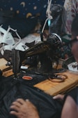 A vintage sewing machine is positioned on a cluttered wooden table surrounded by various objects like fabric and scissors. The background features a starry patterned fabric, enhancing the cozy, workshop atmosphere.