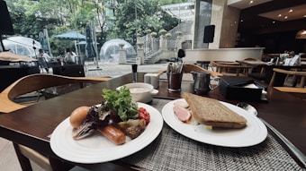 A dining table is set with two plates of food. One plate contains a boiled egg, sausage, leafy greens, grilled vegetables, and cherry tomatoes. The other plate holds a sandwich with slices of ham and lettuce. In the background, glass igloo-style dining pods are visible within a lush garden setting. The ambiance suggests a modern, sophisticated dining area with ample natural light.