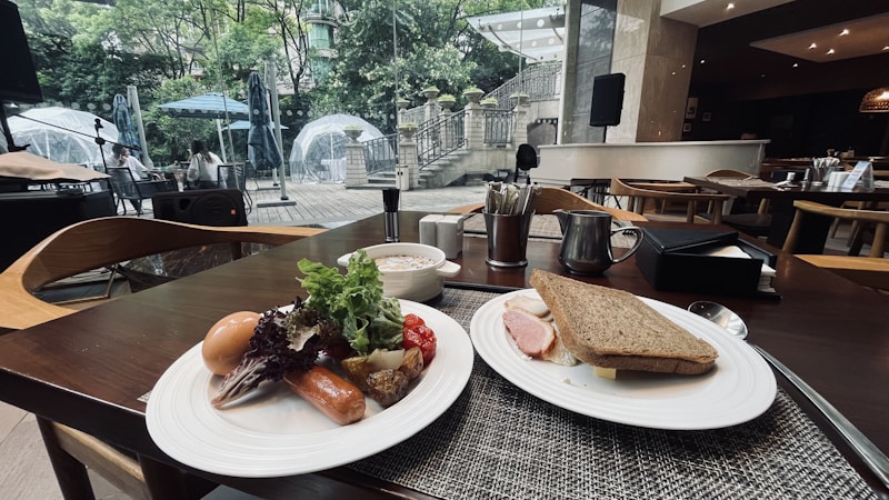 A dining table is set with two plates of food. One plate contains a boiled egg, sausage, leafy greens, grilled vegetables, and cherry tomatoes. The other plate holds a sandwich with slices of ham and lettuce. In the background, glass igloo-style dining pods are visible within a lush garden setting. The ambiance suggests a modern, sophisticated dining area with ample natural light.