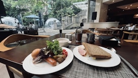 A dining table is set with two plates of food. One plate contains a boiled egg, sausage, leafy greens, grilled vegetables, and cherry tomatoes. The other plate holds a sandwich with slices of ham and lettuce. In the background, glass igloo-style dining pods are visible within a lush garden setting. The ambiance suggests a modern, sophisticated dining area with ample natural light.