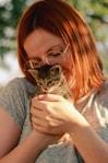 A person with red hair and glasses is gently holding a small tabby kitten in their hands. The scene is lit with natural sunlight, and the background has a soft bokeh effect with greenery.