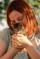 A person with red hair and glasses is gently holding a small tabby kitten in their hands. The scene is lit with natural sunlight, and the background has a soft bokeh effect with greenery.