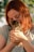 A person with red hair and glasses is gently holding a small tabby kitten in their hands. The scene is lit with natural sunlight, and the background has a soft bokeh effect with greenery.