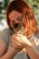 A person with red hair and glasses is gently holding a small tabby kitten in their hands. The scene is lit with natural sunlight, and the background has a soft bokeh effect with greenery.