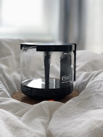 A coffee carafe with the brand 'Technivorm Moccamaster' is resting on a cork mat. The carafe has condensation on its surface, suggesting recent use. The setting seems cozy, with the carafe placed on soft, white fabric.