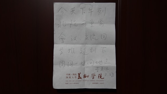 A handwritten note in Chinese, placed against a dark background, possibly a door. The paper includes printed information with red text at the bottom, indicating an association with the Fine Art Academy of Hunan Normal University.