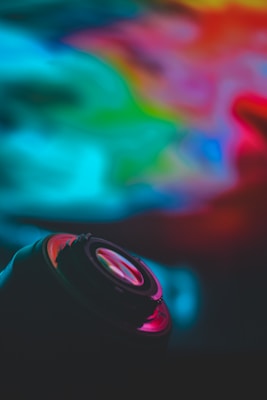 A camera lens is positioned in the foreground, surrounded by vibrant, abstract swirls of color blending into each other. The image has a soft focus and a rich, dark atmosphere.