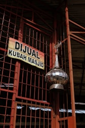 A large metallic dome tilts at the foreground, attached to a structure with orange metal beams and wire mesh. A sign, predominantly yellow with bold black lettering, hangs on the wire mesh, advertising a mosque dome for sale in Indonesian.