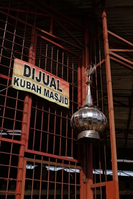 A large metallic dome tilts at the foreground, attached to a structure with orange metal beams and wire mesh. A sign, predominantly yellow with bold black lettering, hangs on the wire mesh, advertising a mosque dome for sale in Indonesian.