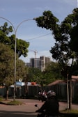 A street view featuring a partially constructed high-rise building in the background, with a crane visible above it. The foreground includes lush green trees, a road with motorcyclists wearing helmets, and a construction site shielded by fencing. A sign with the text 'TK Akbar' is visible near the entrance to the construction area.