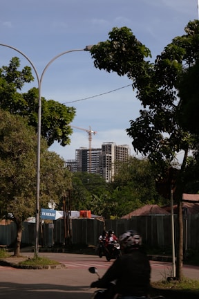 A street view featuring a partially constructed high-rise building in the background, with a crane visible above it. The foreground includes lush green trees, a road with motorcyclists wearing helmets, and a construction site shielded by fencing. A sign with the text 'TK Akbar' is visible near the entrance to the construction area.
