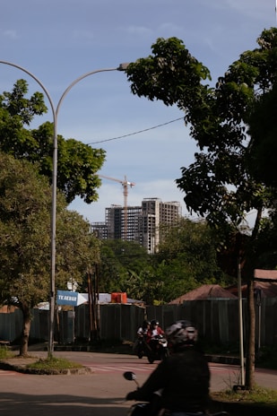 A street view featuring a partially constructed high-rise building in the background, with a crane visible above it. The foreground includes lush green trees, a road with motorcyclists wearing helmets, and a construction site shielded by fencing. A sign with the text 'TK Akbar' is visible near the entrance to the construction area.