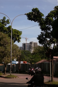 A street view featuring a partially constructed high-rise building in the background, with a crane visible above it. The foreground includes lush green trees, a road with motorcyclists wearing helmets, and a construction site shielded by fencing. A sign with the text 'TK Akbar' is visible near the entrance to the construction area.