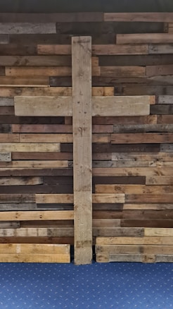 A large wooden cross is mounted on a wall made of variously colored, horizontally laid wooden planks. The planks have a rustic and aged appearance, creating a textured and warm background. Below the cross and wall is a blue carpeted floor.