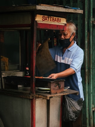 A street vendor wearing a mask is cooking food on a small cart. The sign on the cart reads 'Sate Babi' which indicates that pork satay is being sold. The vendor is using a traditional charcoal grill. The cart looks worn, suggesting long-term use, and is parked in an urban setting with metallic backgrounds.
