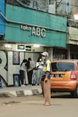 A street scene featuring a teal-colored storefront sign with 'toko ABC' written on it. A group of people stands near the entrance, one of whom is wearing a beige headscarf and another in a black hooded jacket. An orange car is parked on the side of the road, and a man wearing a blue and yellow shirt, brown pants, and a hat stands in front of it, looking off to the side.
