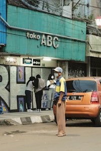 A street scene featuring a teal-colored storefront sign with 'toko ABC' written on it. A group of people stands near the entrance, one of whom is wearing a beige headscarf and another in a black hooded jacket. An orange car is parked on the side of the road, and a man wearing a blue and yellow shirt, brown pants, and a hat stands in front of it, looking off to the side.