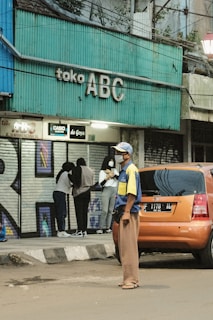 A street scene featuring a teal-colored storefront sign with 'toko ABC' written on it. A group of people stands near the entrance, one of whom is wearing a beige headscarf and another in a black hooded jacket. An orange car is parked on the side of the road, and a man wearing a blue and yellow shirt, brown pants, and a hat stands in front of it, looking off to the side.