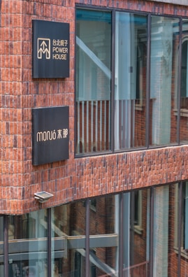 A modern building facade with large glass windows framed by reddish-brown bricks. Two black signs with gold inscriptions and symbols are mounted on the wall. Reflections of nearby architecture can be seen in the windows.