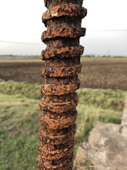 A close-up of a rusted piece of rebar, with significant corrosion and chipping, is captured in front of an agricultural field. The corroded surface of the rebar displays a distinctive texture and rich orange-brown color. The background features a plowed farm with a grassy area in the foreground, creating a strong contrast with the rugged surface of the rebar.