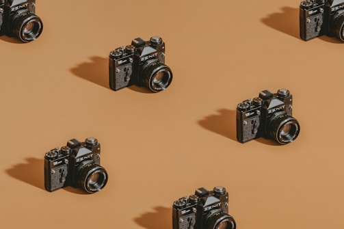 Vintage cameras are neatly arranged in a repeating diagonal pattern on a warm brown background. The lighting creates shadows, adding depth and texture to the scene.
