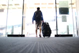 A person is walking through a glass entrance door, pulling a large travel suitcase with one hand. The individual is wearing casual clothing, including shorts and a backpack. The setting appears to be an indoor area, potentially an airport or terminal, indicated by the design of the sliding doors and signage.