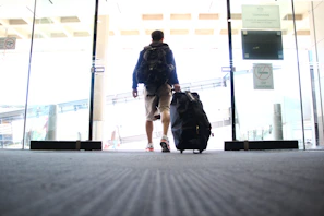 A person is walking through a glass entrance door, pulling a large travel suitcase with one hand. The individual is wearing casual clothing, including shorts and a backpack. The setting appears to be an indoor area, potentially an airport or terminal, indicated by the design of the sliding doors and signage.