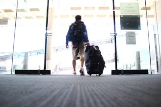A person is walking through a glass entrance door, pulling a large travel suitcase with one hand. The individual is wearing casual clothing, including shorts and a backpack. The setting appears to be an indoor area, potentially an airport or terminal, indicated by the design of the sliding doors and signage.