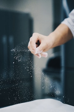 A hand is sprinkling a fine white powder, likely flour, over a surface. The background is blurred, emphasizing the motion and texture of the powder in mid-air.