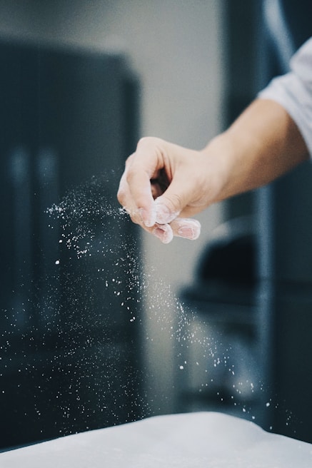 A hand is sprinkling a fine white powder, likely flour, over a surface. The background is blurred, emphasizing the motion and texture of the powder in mid-air.