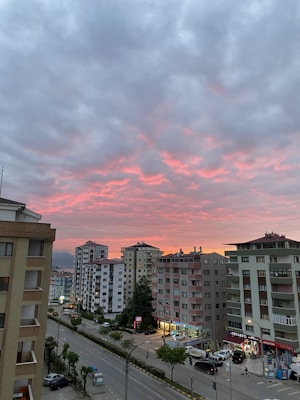 A vibrant sunset with pink and orange hues in the sky, casting a warm glow over a cityscape. Several multi-story apartment buildings line the street below, with cars parked alongside and small shops with illuminated signs at the ground level. Trees are sparsely scattered along the sidewalk.