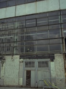 A large windowed facade of a building with reflective glass panes. The reflection shows another building in a similar industrial style. The lower part of the structure consists of worn, peeling white bricks and a set of double doors that appear old and weathered. A metal railing with yellow caution tape runs along the front.