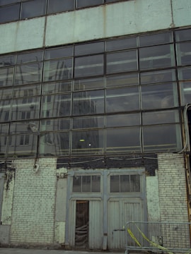 A large windowed facade of a building with reflective glass panes. The reflection shows another building in a similar industrial style. The lower part of the structure consists of worn, peeling white bricks and a set of double doors that appear old and weathered. A metal railing with yellow caution tape runs along the front.