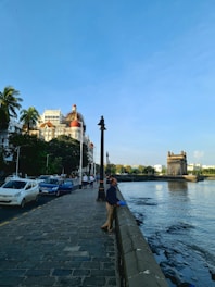 A scenic waterfront with a historic hotel featuring red domes. The foreground has a walkway lined with streetlamps and people strolling. Cars are parked along the pavement. A stone gateway structure stands prominently in the water near the shore, with distant buildings and trees in the background under a clear blue sky.